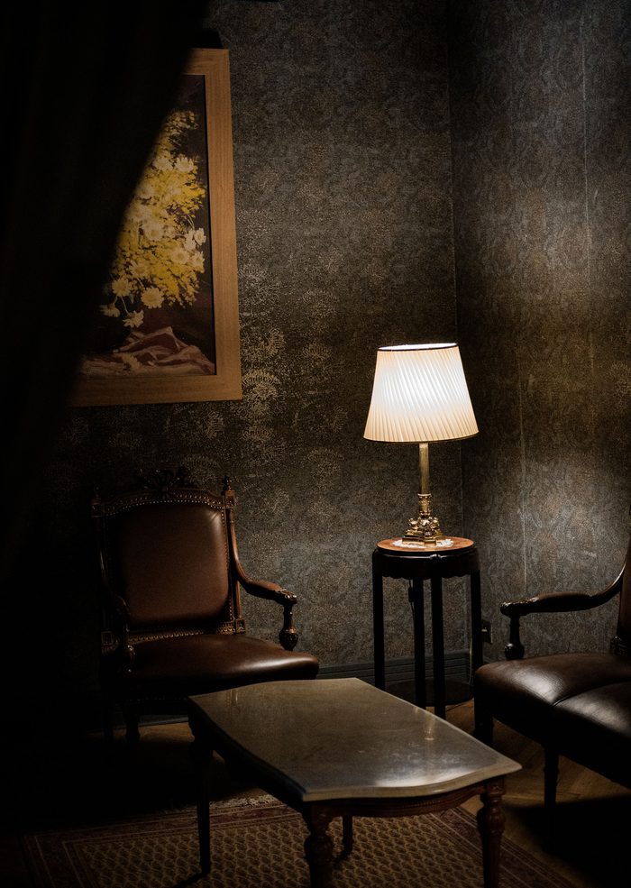 Dimly lit sitting room with a dark patterned wallpaper, leather armchairs, a glass coffee table and a pleated table lamp