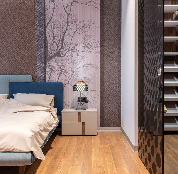 Bedroom with a misty lilac tree-branch mural behind a blue upholstered bed and a sculptural lamp