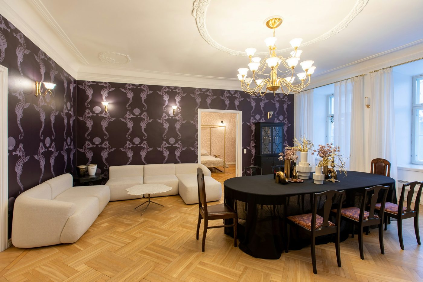 Elegant living and dining room with a dramatic aubergine seahorse-pattern feature wall, a brass chandelier and cream modular sofas