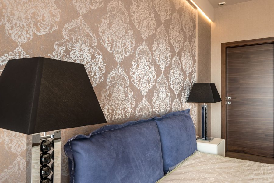 Bedroom wall covered in a silver damask wallpaper behind a blue velvet headboard and a black-shaded lamp