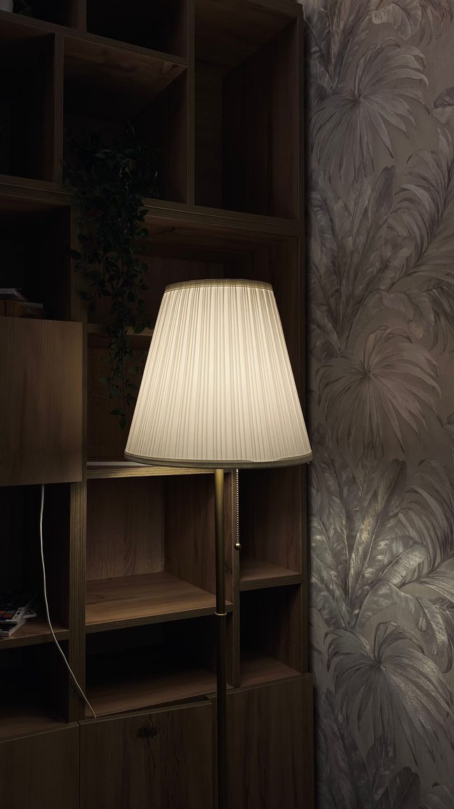 Pleated lamp glowing in front of a dark, softly sheened leaf-pattern wallpaper beside built-in wooden shelving